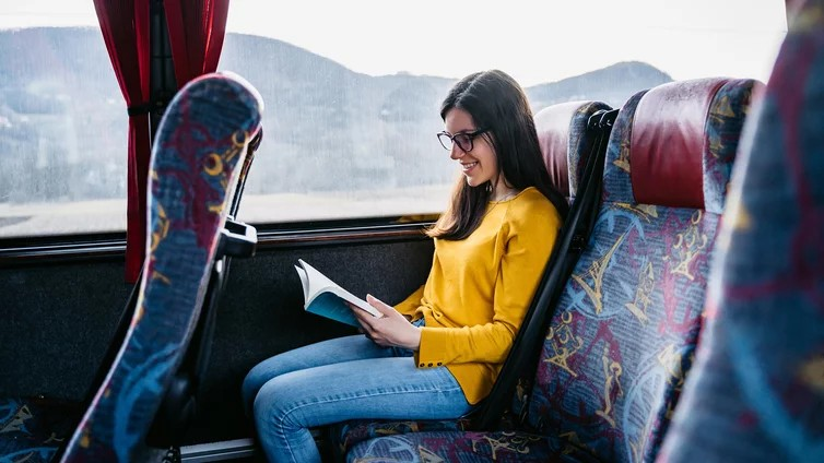 Fotografie einer Frau mit gelbem Pullover und blauer Jeans, die in einem Zug am Fenster sitzend ein Buch liest. Im Hintergrund sind durch das Fenster unscharfe Berglandschaften zu sehen, während bunte Sitzbezüge mit abstrakten Mustern den Innenraum desZug
